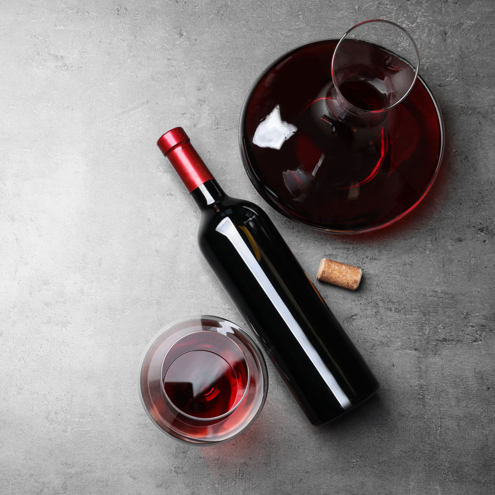 Μία καράφα κρασιού με φαρδιά βάση γεμάτη με κρασί με ένα ξαπλωμένο μπουκάλι και ένα ποτήρι κρασιού σε γκρι φόντο